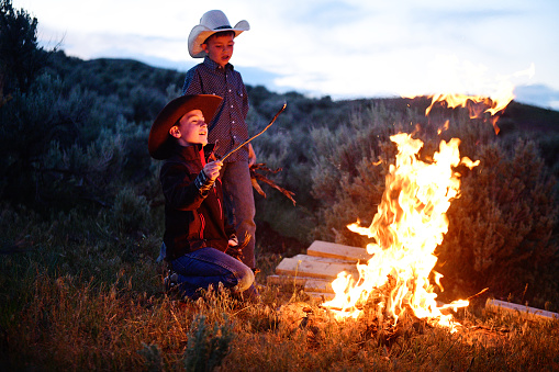 This is a color photograph of two little cowboys  watching their camp fire burning at dusk in rural Utah, USA.