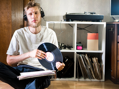 young man sitting in cozy living room holding the vinyl record disk near the records rack on the floor