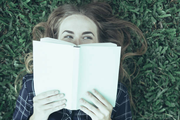 istock Smiling Girl Lying on Grass and Hiding Behind Book 1012420080