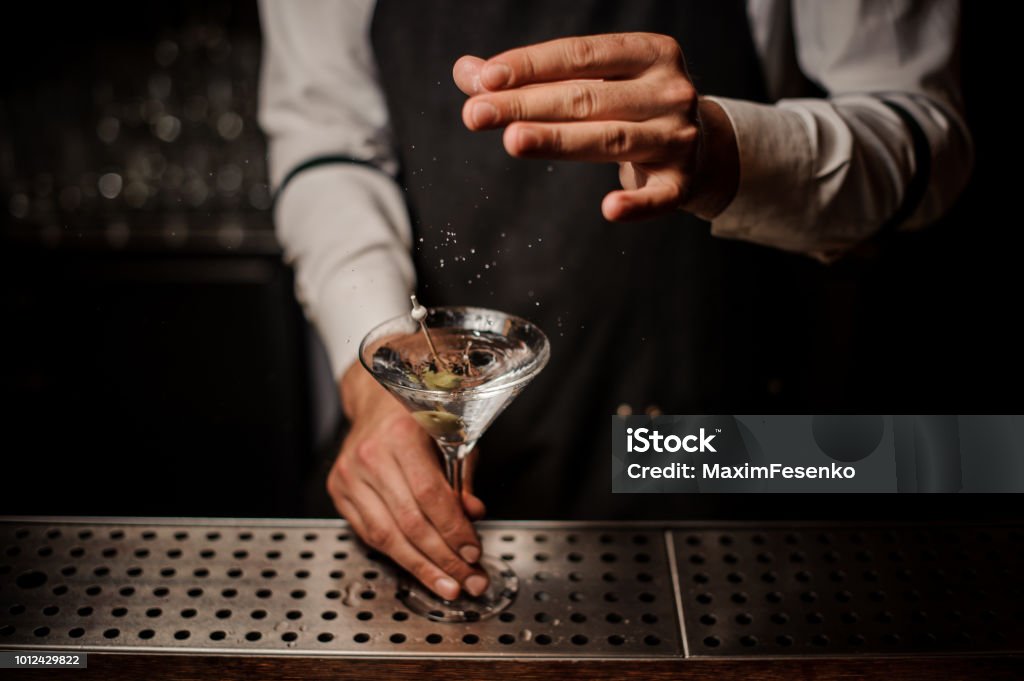 Barman adding salt into a strong martini cocktail Barman adding salt into a strong martini cocktail decorated with a green olive on the bar Martini Stock Photo