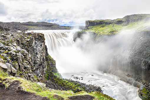 Icelandic Dettifoss waterfall, Iceland, largest volume in Europe, gray grey water, rocky cliff, rocks, soil, green grass, water flowing mist spraying, people walking