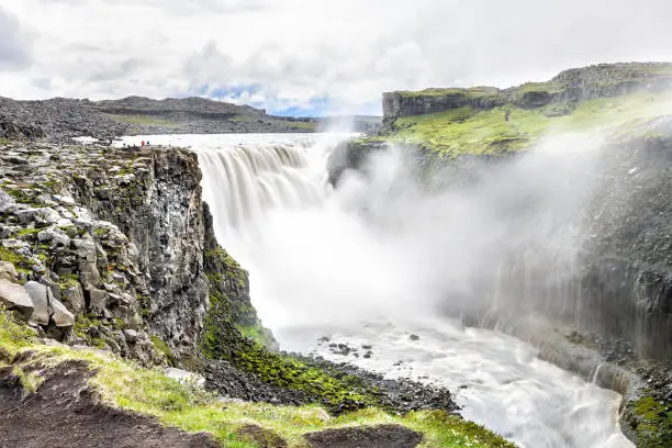 Photo of Icelandic Dettifoss waterfall, Iceland, largest volume in Europe, gray grey water, rocky cliff, rocks, soil, green grass, water flowing mist spraying, people walking