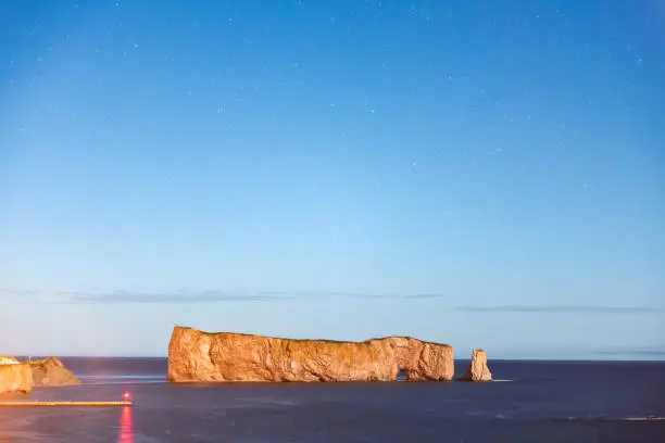 Famous Rocher Perce rock in Gaspe Peninsula, Quebec, Gaspesie region, Canada at dark night starry sky, stars at Saint Lawrence gulf, wharf, dock, pier, lighthouse, reflection