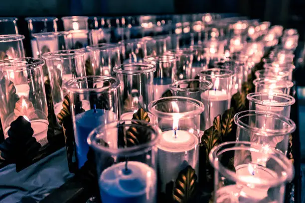 Closeup side pattern of many white, blue religious votive candles with yellow, purple flame in church or cathedral