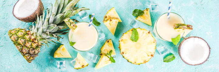 Refreshing summer drink, homemade pina colada cocktail, on a light blue background, with pieces of pineapple, coconut, ice and mint leaves, copy space top view banner