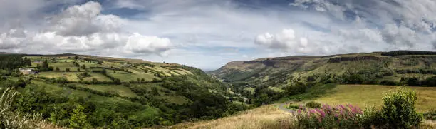 a sunny panoramic summer view of the valley at Glenariff, County Antrim, Northern Ireland, with rolling hills of agricultural fields and a grassy foreground, under a broken sky