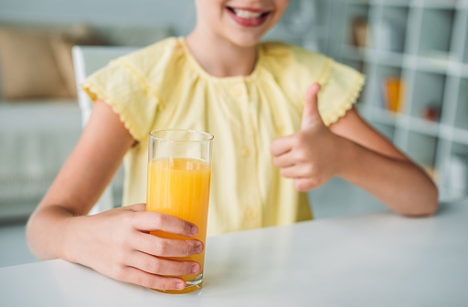 cropped shot of smiling little child with glass of orange juice showing thumb up