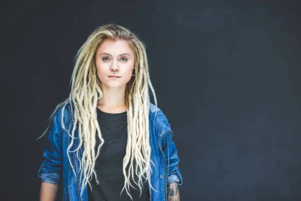 Portrait of young woman with blond dreadlocks and piercing standing against dark background