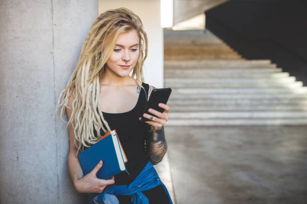 Woman with dreadlocks using phone at university Portrait of young woman with dreadlocks and piercing holding books and texting on smart phone at university education student mobile phone university stock pictures, royalty-free photos & images