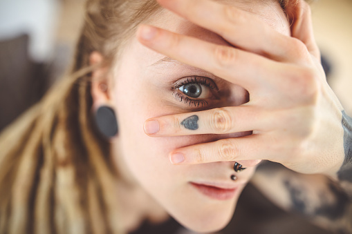 Close up portrait of young woman looking through her fingers, focus on grey eyes on caucasian woman, with tattooed hand.