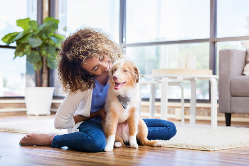 A young woman sits on the floor of her living room with her new puppy and embraces him as she leans forward and smiles.