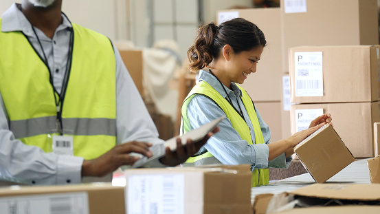 Confident male and female distribution warehouse workers prepare to send out packages to customers. A female employee is placing a mailing label on the package. A male employee is using a digital table tin the foreground.