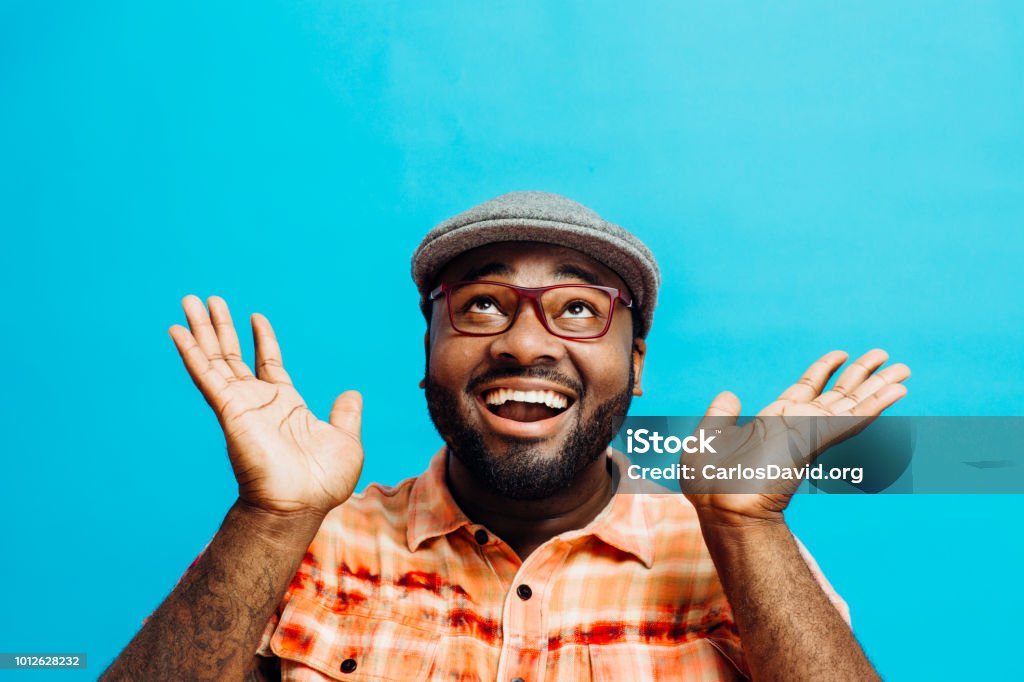 It's incredible! Portrait of a happy and excited man looking up. It's incredible! Portrait of a happy and excited man looking up with mouth open and both arms up, in front of a blue background People Stock Photo