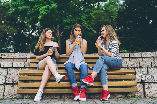 Group of teenage girls sitting on a bench and drinking orange juice, downtown.