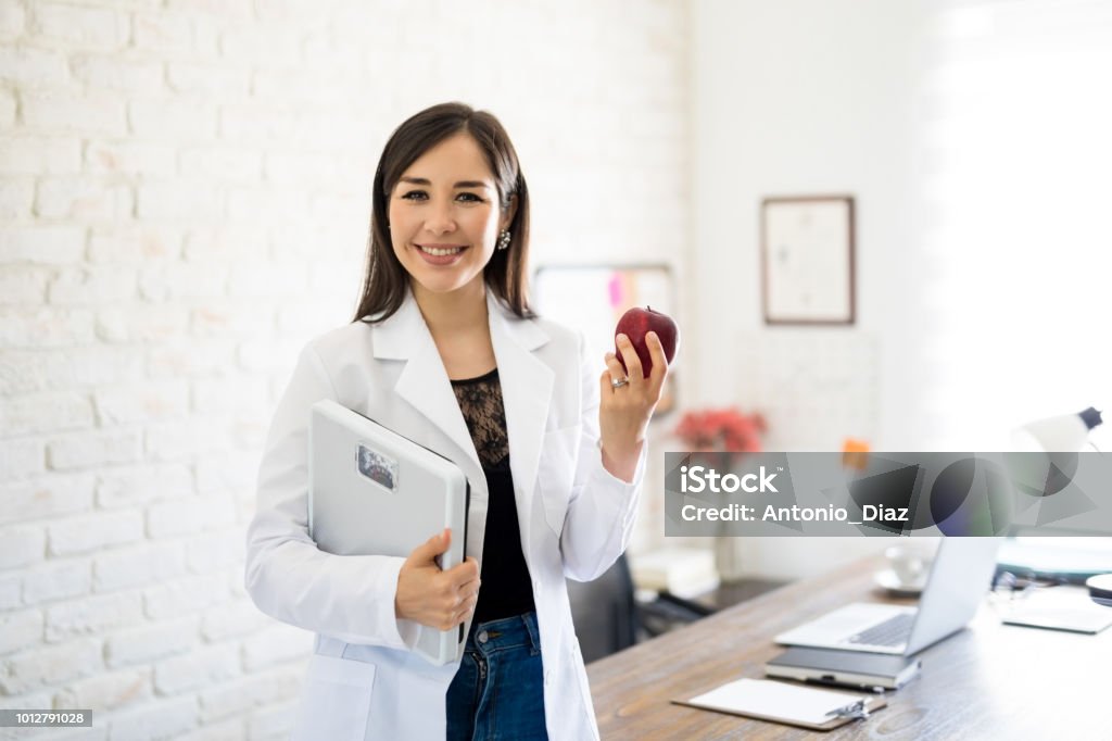 Dietitian with weight scale and apple Portrait of young smiling female nutritionist holding weight scale and apple in the consultation room Nutritionist Stock Photo