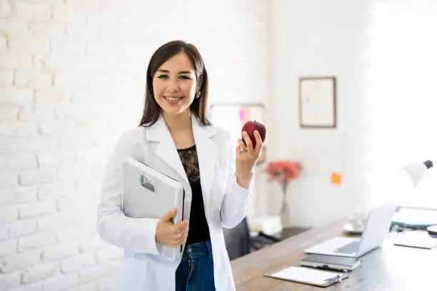 Portrait of young smiling female nutritionist holding weight scale and apple in the consultation room