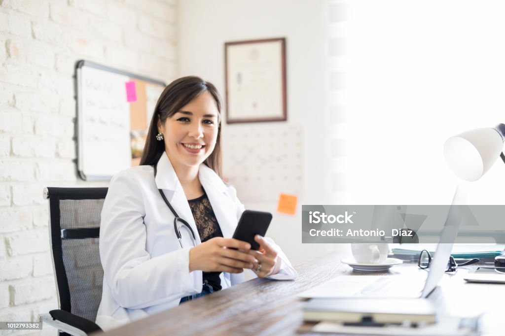 Happy young doctor with phone at office Portrait of cheerful female doctor sitting at her desk and using mobile phone Doctor Stock Photo