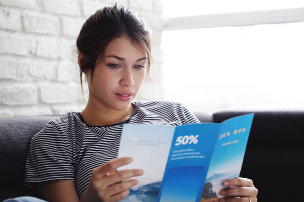 Woman Reading Travel Flye On Sofa At Home Young hispanic woman laying on couch at home, holding a travel flyer and planning next trip. Copy space on window. woman flyers stock pictures, royalty-free photos & images