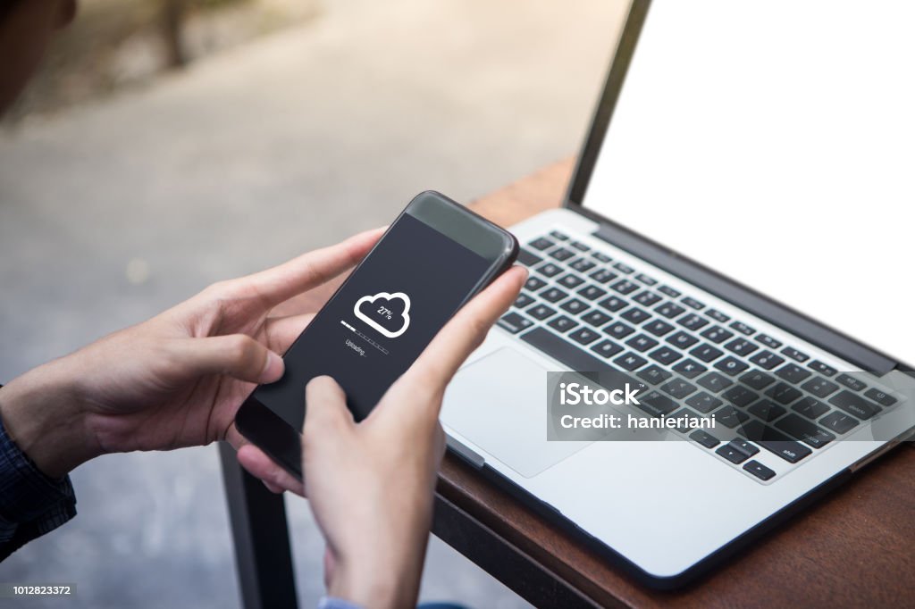 Während der Arbeit am Laptop mann tut Cloud hochladen auf seinem Smartphone auf Terrasse - Lizenzfrei Herunterladen Stock-Foto