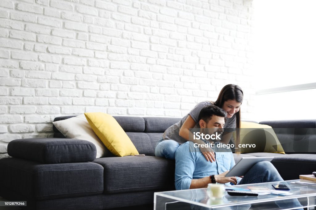 Girl Massaging Boyfriend On Sofa At Home Young hispanic couple sitting on couch at home, using a tablet PC for Internet and social media. The girl is giving a massage to her boyfriend. Copy space Couple - Relationship Stock Photo