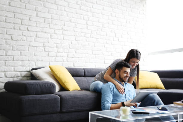 Girl Massaging Boyfriend On Sofa At Home Young hispanic couple sitting on couch at home, using a tablet PC for Internet and social media. The girl is giving a massage to her boyfriend. Copy space lifestyle couple stock pictures, royalty-free photos & images
