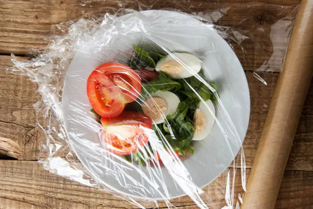 Roll of wrapping plastic stretch film over plate of vegetabes. Close-up isolated on wooden background. No plastic concept.