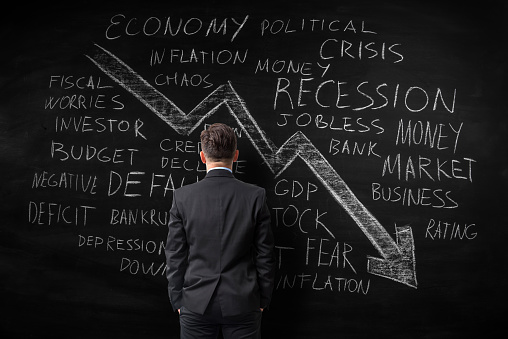 Economic crisis and related business therms word cloud written with chalk on black board. There is a big arrow chart moving down. A businessman seen from the back is looking at the blackboard