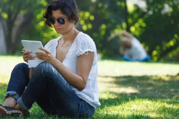 The brunette girl in the park is reading a record. A concept of calm mood and relaxation in a park on the green grass. A middle-aged woman in blue jeans.