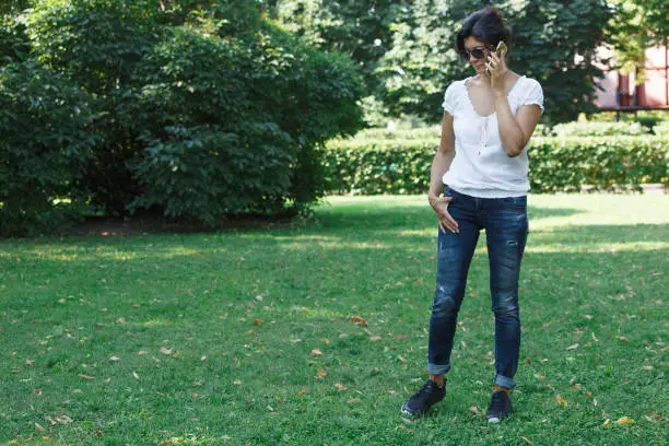The brunette girl in the park is calling by mobile phone. A concept of calm mood and relaxation in a park on the green grass. A middle-aged woman in blue jeans.