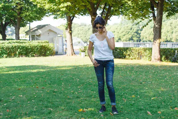The brunette girl in the park is calling by mobile phone. A concept of calm mood and relaxation in a park on the green grass. A middle-aged woman in blue jeans.