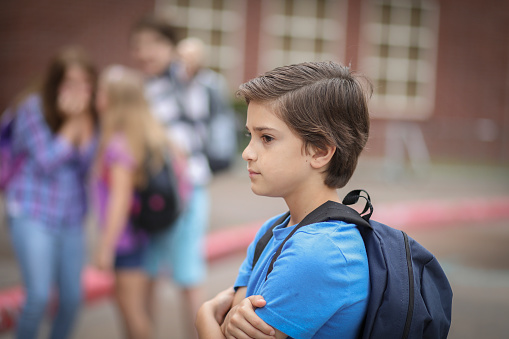 Elementary age boy being bullied outside the elementary school building,  Other students in background laugh at boy in background.