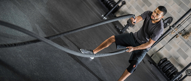 Panoramic photo of a man of African ethnicity swinging battle ropes in a gym.