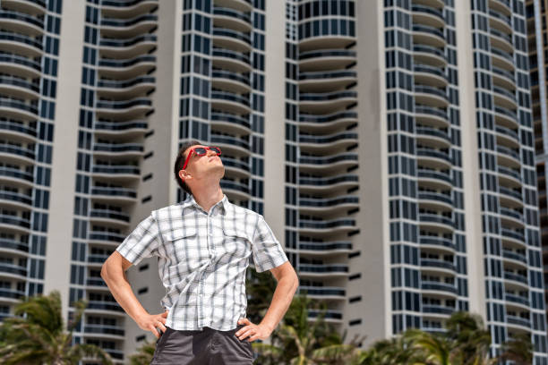 istock Young handsome attractive man hipster millennial looking up on beach during sunny day with red sunglasses in Miami, Florida with hands on hips, apartment condo skyscraper 1013463736