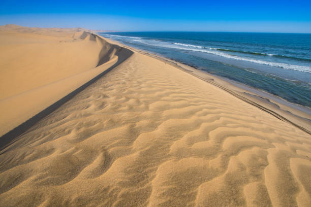 Rolling waves kiss the base of towering sand dunes along the coast of Sandwich Harbour, Namibia. This surreal scene captures the raw beauty where the Namib Desert collides with the Atlantic Ocean in a seamless meeting of land and water.