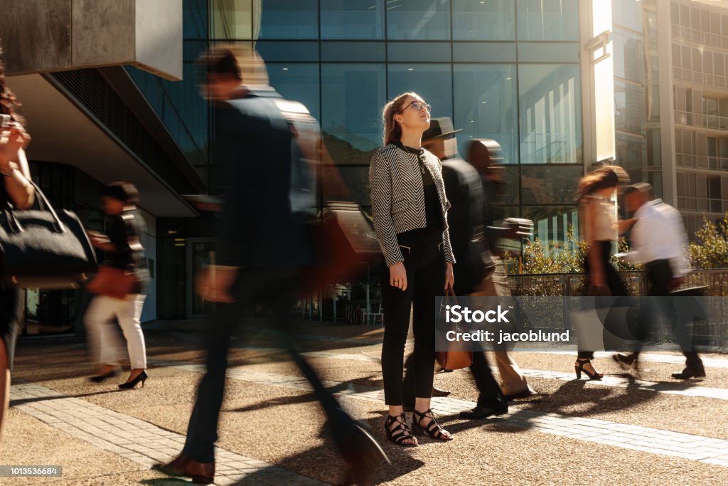 Businesswoman standing still on a busy street Woman standing amidst a busy office going crowd hooked to their mobile phones. Businesswoman holding her hand bag standing still on a busy street with people walking past her using mobile phones. People Stock Photo