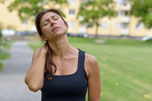 Middle aged woman out jogging suffers a muscle injury standing holding her neck while grimacing in pain, close up upper body view