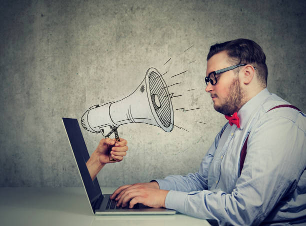 istock man sitting at table working on computer screaming with a megaphone poking out from a laptop screen 1013559074