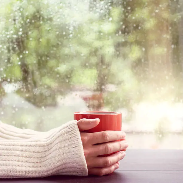 man in a light jumper, holds in hands red mug on a background of a wet window after the rain