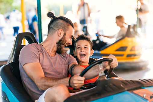 Photo of Caucasian young hipster Father and his 5 years old son having a ride in the bumper car at the amusement park during summer day. Happy family leisure in holiday weekend