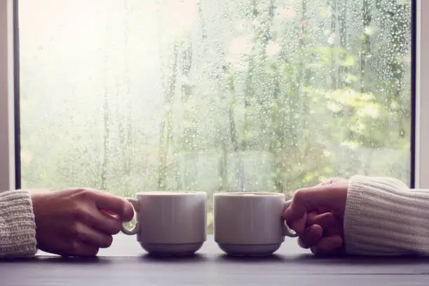 two white cups in hands on a background of wet window with raindrops