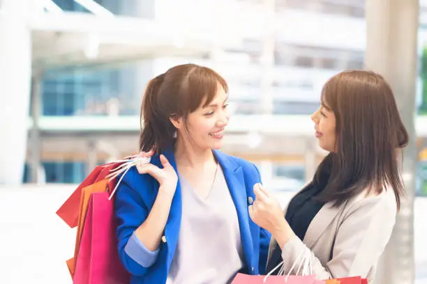 Asian women holding colorful paper shopping bags in both hands and chatting together while walking outside departmentstore