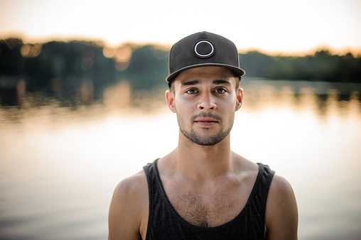 Attractive young man in the dark cap looking at camera standing on the background of a lake