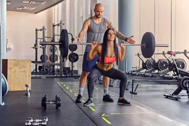 Sporty shaved head male coach assisting to a female doing squats with a barbell.