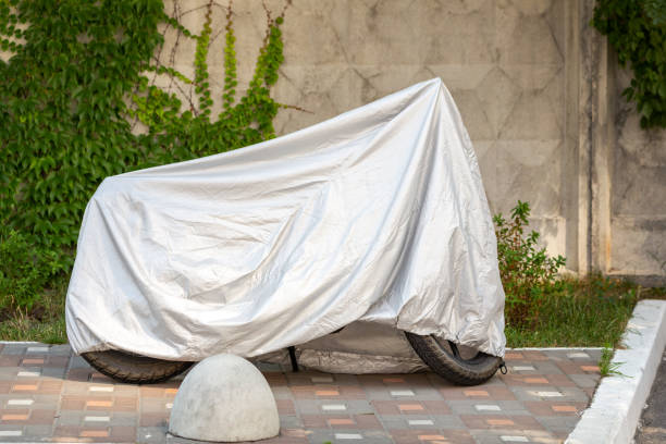 istock Motorcycle covered with grey protective jacket . Scooter with fabric shield parked at pavement near concrete wall. Waterproof parking outdoors during travel 1013697962