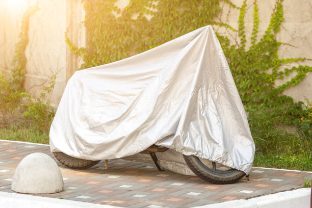 istock Motorcycle covered with grey protective jacket . Scooter with fabric shield parked at pavement near concrete wall. Waterproof parking outdoors during travel 1013697978