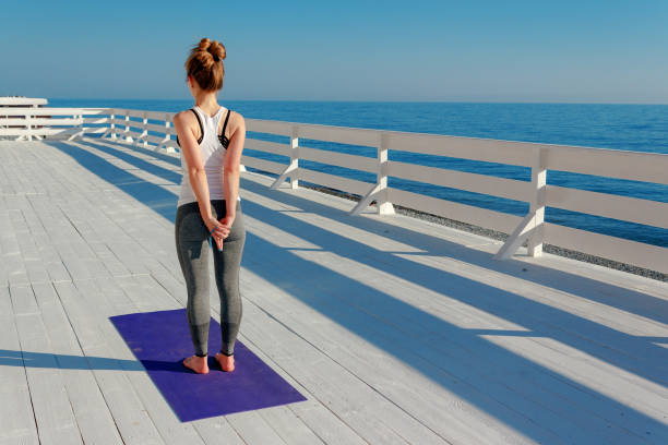 Young slim woman stretching outdoors at white wooden seafront. Girl standing on violet yoga mat with hands back in mudra. Exercise for shoulder blades Young slim woman stretching outdoors at white wooden seafront. Girl standing on violet yoga mat with hands back in mudra. Exercise for shoulder blades. scapula stock pictures, royalty-free photos & images