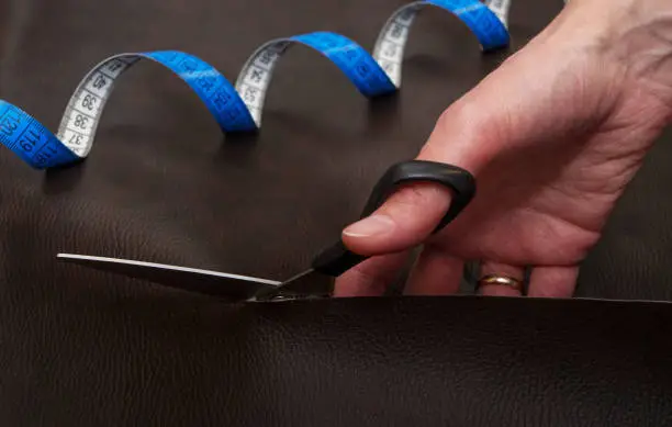 Woman's hand holding black scissors and cutting out leather, blue twisted tape measure on the background. Close-up