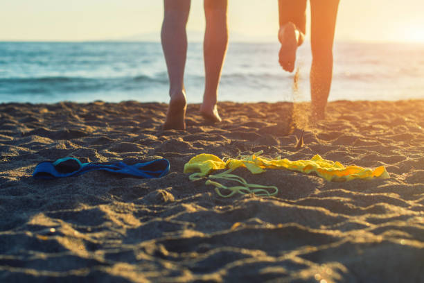 gambe femminili e maschili con bikini e costumi da bagno sulla sabbia su uno sfondo al tramonto. - nudo foto e immagini stock