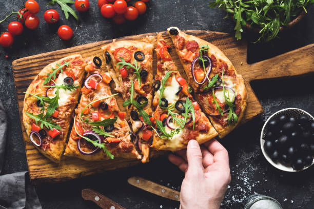 Flatbread pizza garnished with fresh arugula on wooden pizza board, top view Flatbread pizza garnished with fresh arugula on wooden pizza board, top view. Dark stone background. Person picking slice of pizza flatbread stock pictures, royalty-free photos & images