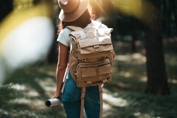 istock Back view on cute young woman with hat, backpack and location map in hand among trees in forest at sunset. Lens flare effect 1013816654